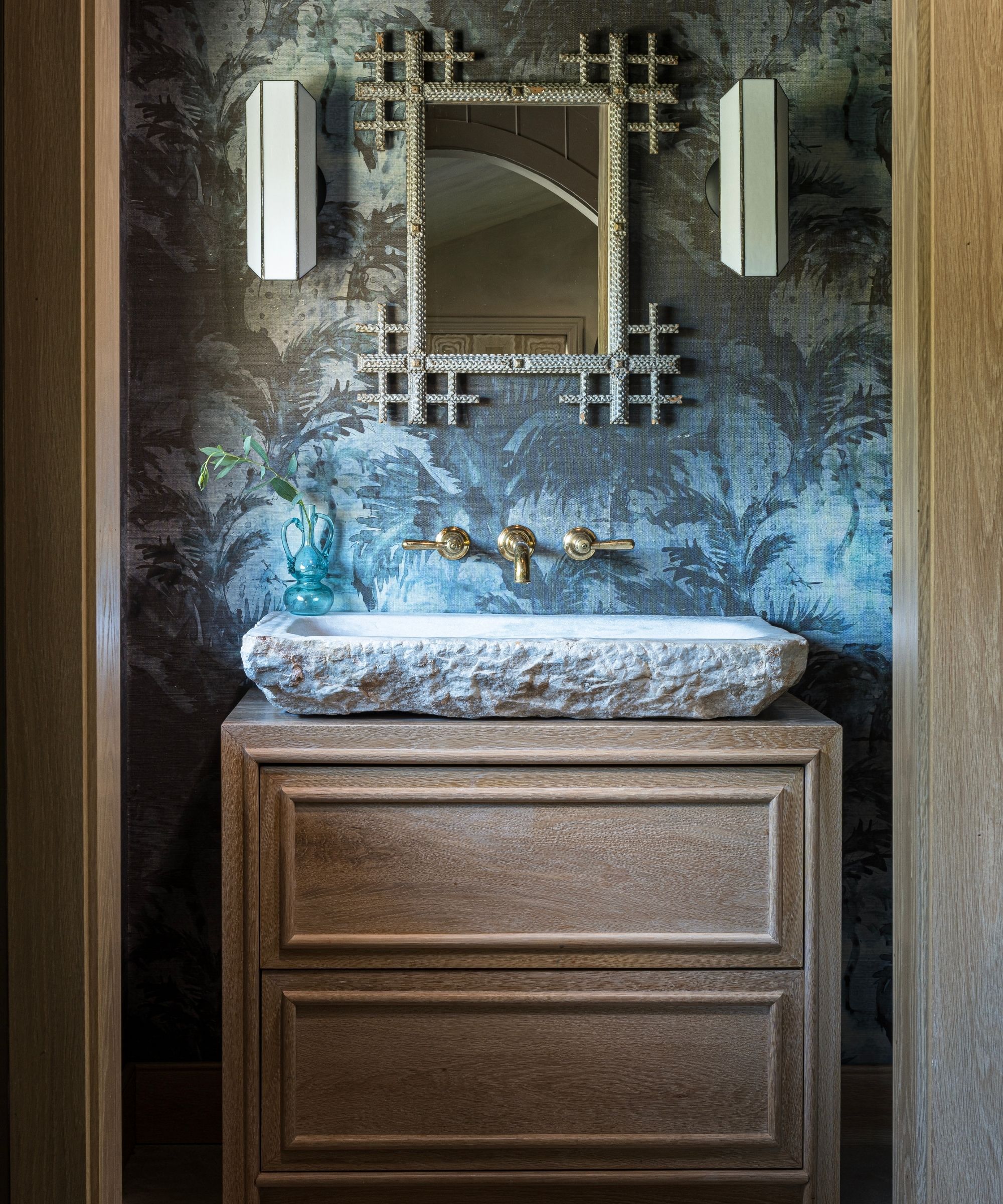 a blue-gray wallpapered leafy bathroom with stone and oak vanity and vintage mirror