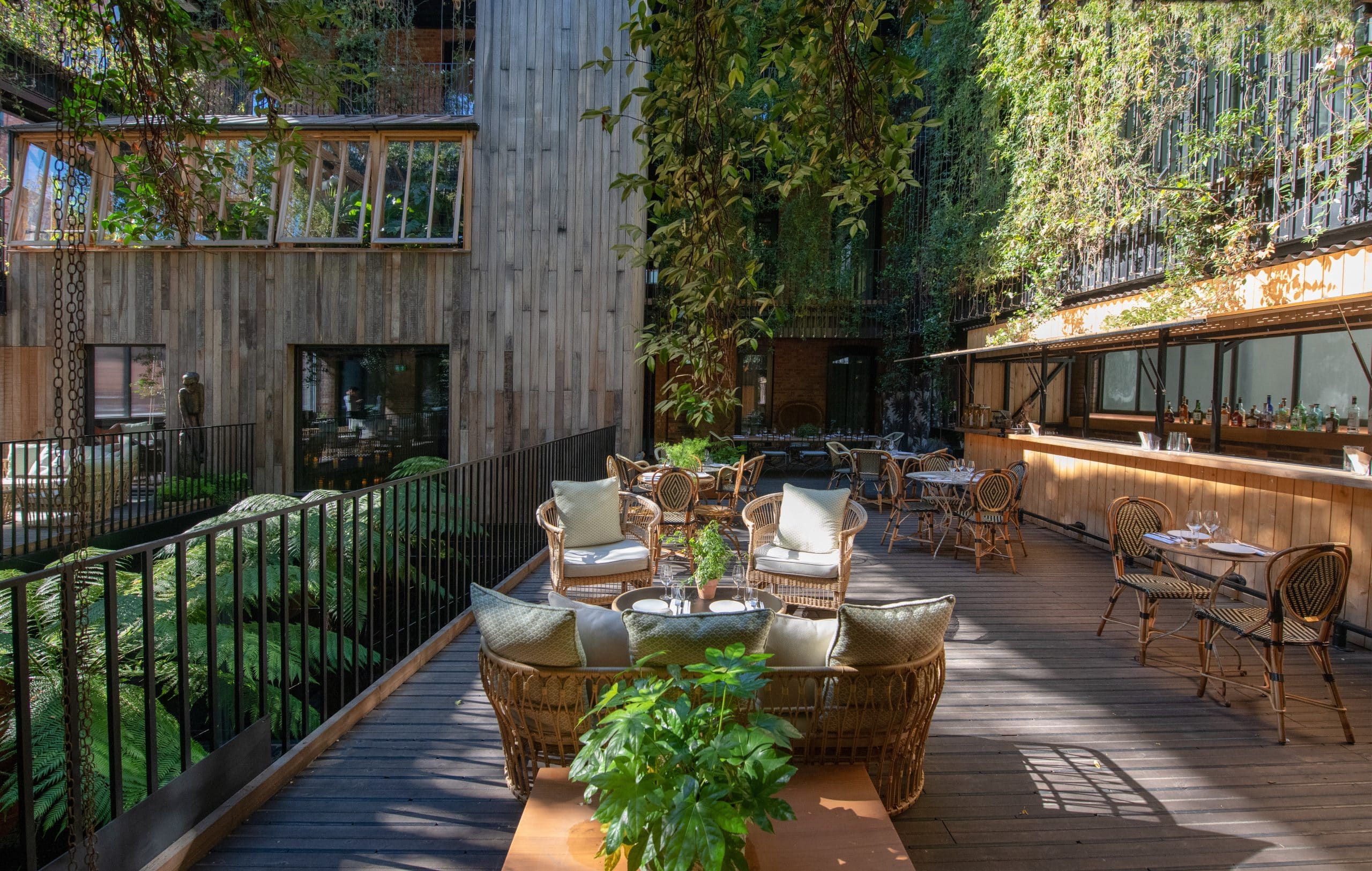 An outdoor terrace with wooden flooring and wood cane chairs and tables. Surrounding the eating area are cascading green vines.