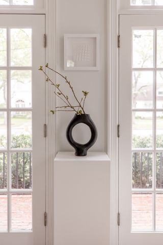 A living room with a black ceramic vase on a white pedestal containing stems of budding leaves