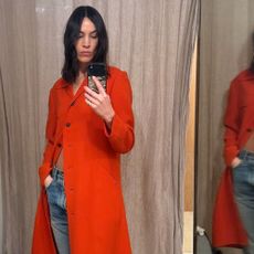 Alexa Chung takes a mirror selfie wearing a tomato red jacket with blue jeans and black ballet flats.