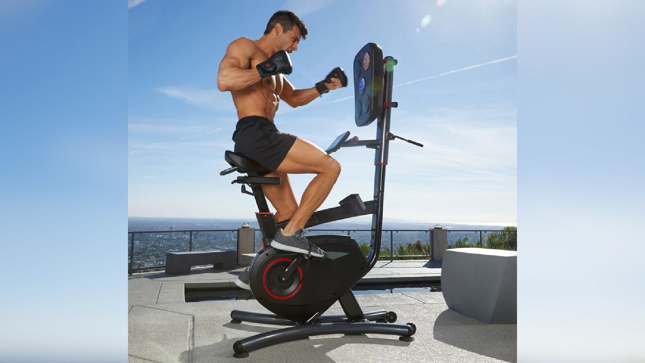 A topless man sits on a CycleBoxer bike while punching at the intergrated pads on a roof garden with blue skys