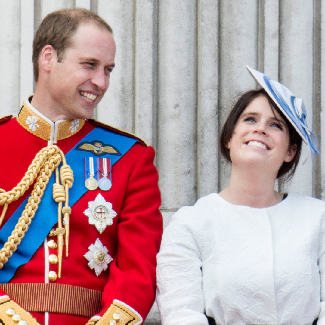 Prince William laughs while wearing red military uniform as he stands next to Princess Eugenie, who is wearing a white dress and a blue and white hat on the Buckingham Palace balcony