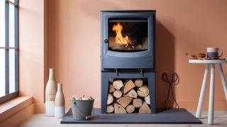 black log burner with log store below installed on black hearth in modern room with pale terracotta walls, small white table, log bucket and floor standing vases