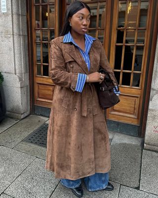 @nnennaechem wears a brown suede trench coat with a stripe shirt, jeans and boots