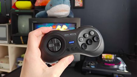 Hand holding 8Bitdo M30 2.4GHz controller in front of desk with Japanese Sega Mega Drive connected to Sony Trinitron CRT TV with BL&Aring;HAJ Ikea shark on top and Golden Axe title on screen.