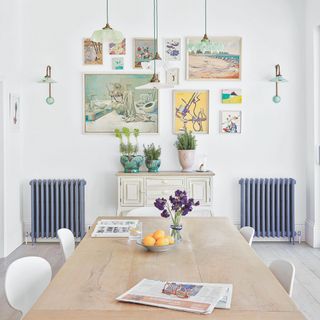 White dining room with wooden dining room table and white chairs, with two blue radiators on the wall and a gallery wall of prints