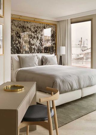 Interior view of a room at The One, Spain featuring light coloured walls, wooden flooring, a rug, a bed with white and grey pillows and linen, a gold framed reflective headboard with a white feather style design, a tall window, white curtains, framed wall art, a lamp, a desk and a chair