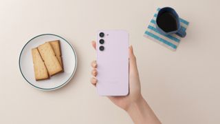 The Samsung Galaxy S23 Plus, held in hand next to a plate of biscuits and a mug of coffee