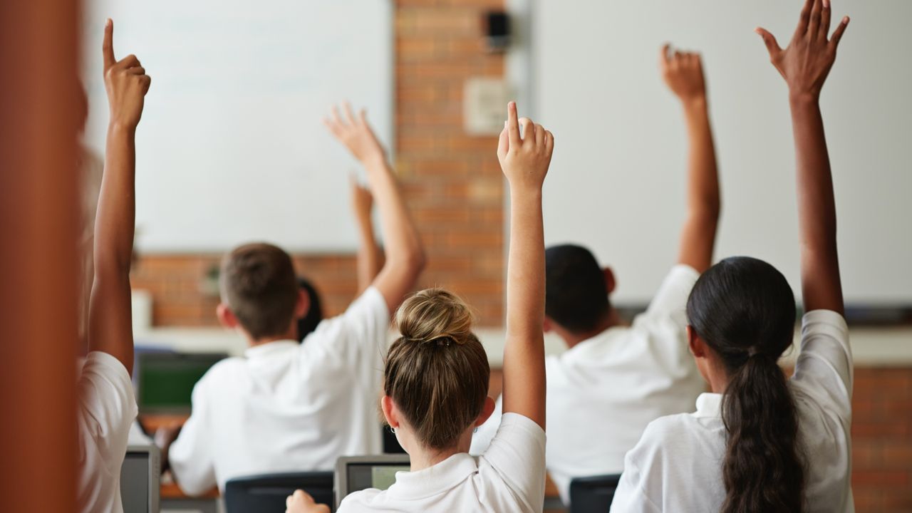 Young girls in a classroom, raising their hands. 