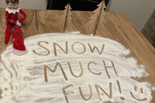 Our writer's Elf sitting on a table surrounded by spilled flour in which someone has spelled out the words 'Snow Much Fun'.