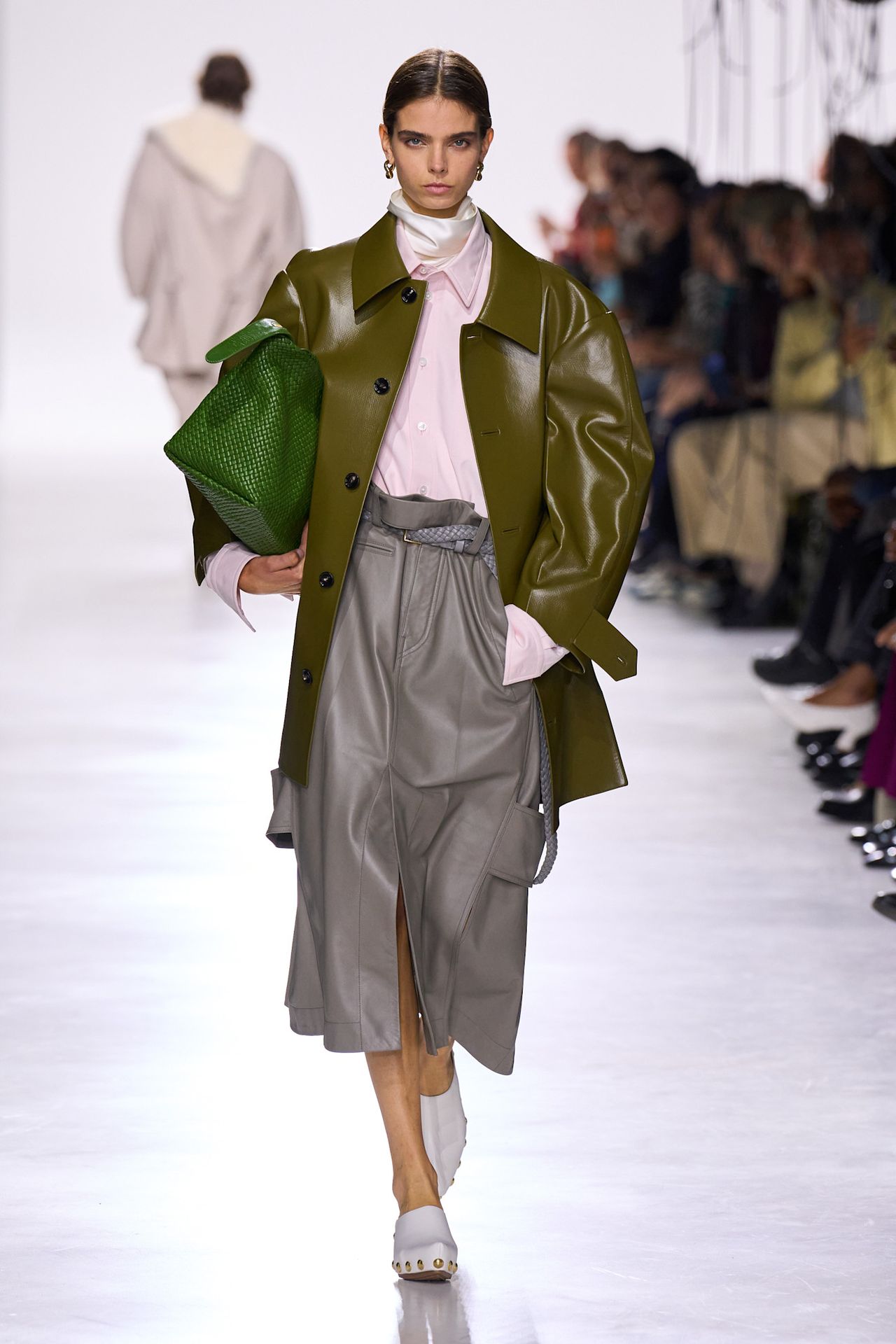 A Bottega Veneta model wearing an olive green leather jacket, a pink button-down shirt with a white turtleneck peaking out of the collar, a gray leather midi skirt, white clog shoes, and a bright green tote bag.
