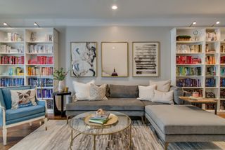 Home library with shelving, neutral L-shaped sofa, blue armchair, wood floor, glass coffee table, neutral patterned rug, neutral wall with artwork