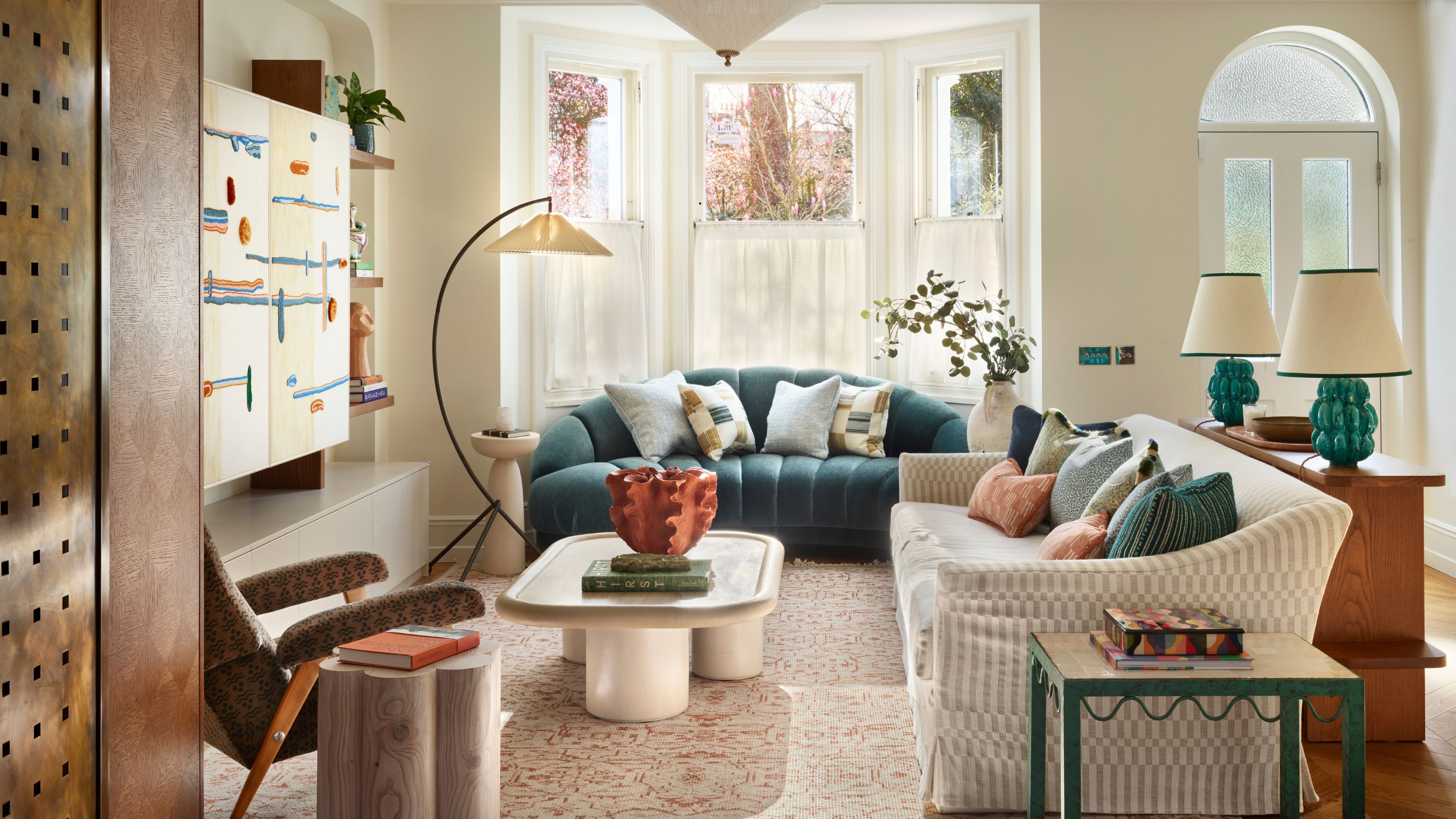 neutral living room with large bay window, a teal velvet sofa with a stone coffee table, large floor lamp, slip covered sofa styled with a table behind it with two teal lamps