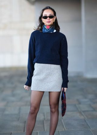 A guest wears sunglasses, a blue and red floral print scarf, a navy blue pullover , a gray ribbed wool mini skirt , black tights, pointed shoes with kitten heels, outside OperaSport, during the Copenhagen Fashion Week AW24 on January 31, 2024 in Copenhagen, Denmark.