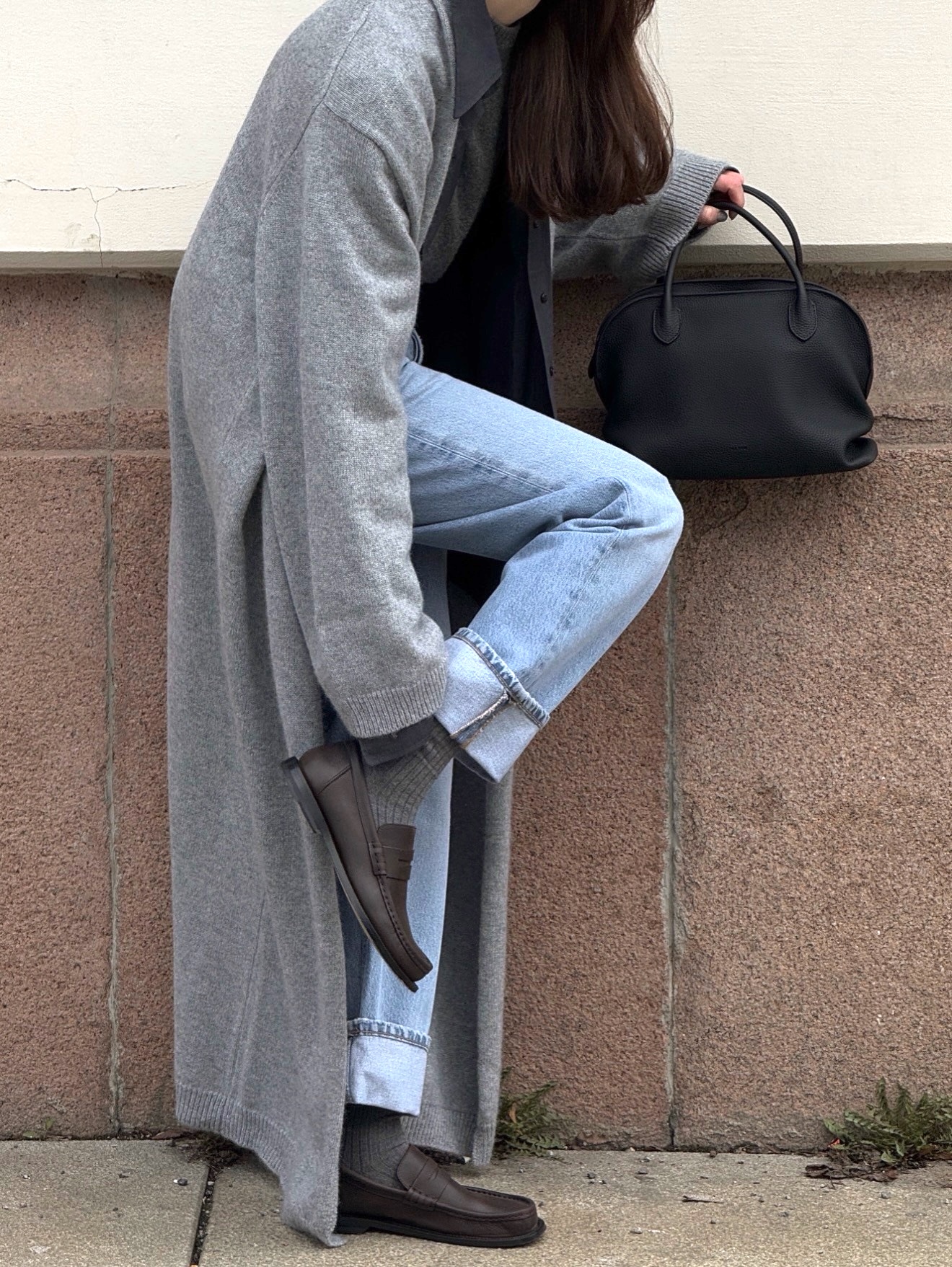Style influencer Annabel Rosendahl poses wearing a maxi gray sweater jacket, a black bag, cuffed jeans, gray socks, and brown loafers.