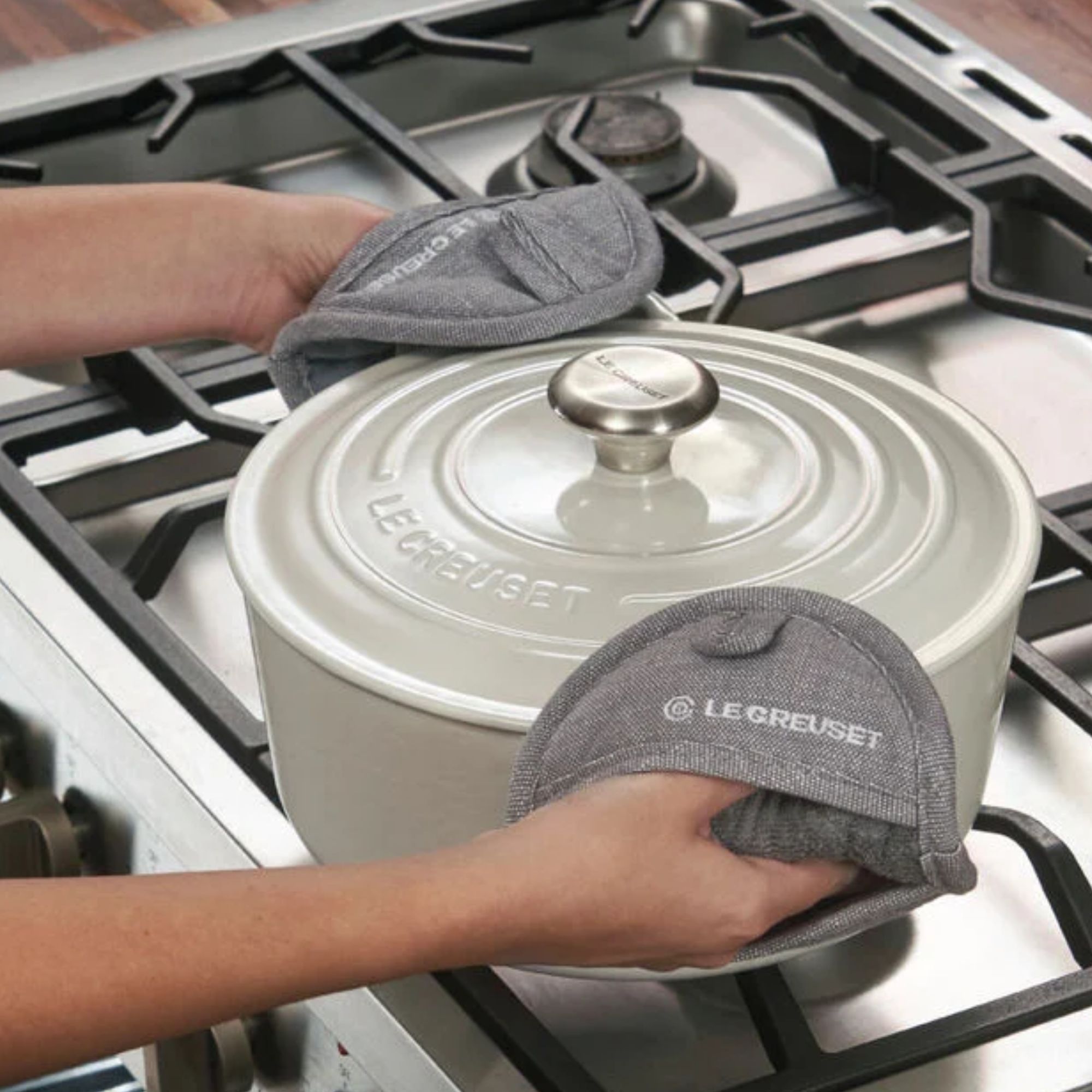 Someone using grey fabric pot holders to lift a grey Dutch oven from a gas stove.