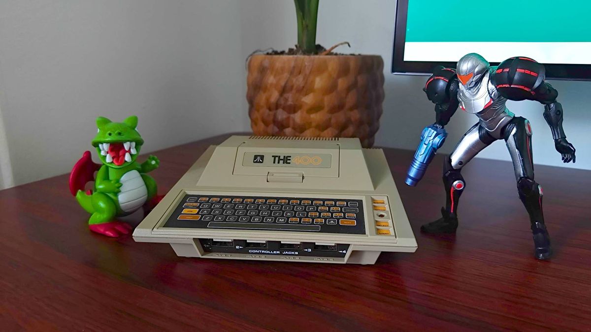 Atari 400 Mini console with Samus figure on right and Dragon from Dig-Dug on left sitting on woodgrain desk