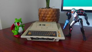Atari 400 Mini console with Samus figure on right and Dragon from Dig-Dug on left sitting on woodgrain desk