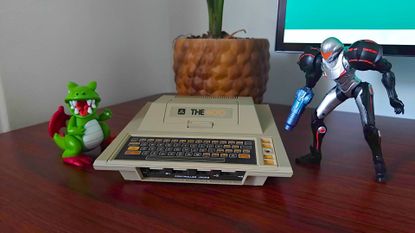 Atari 400 Mini console with Samus figure on right and Dragon from Dig-Dug on left sitting on woodgrain desk