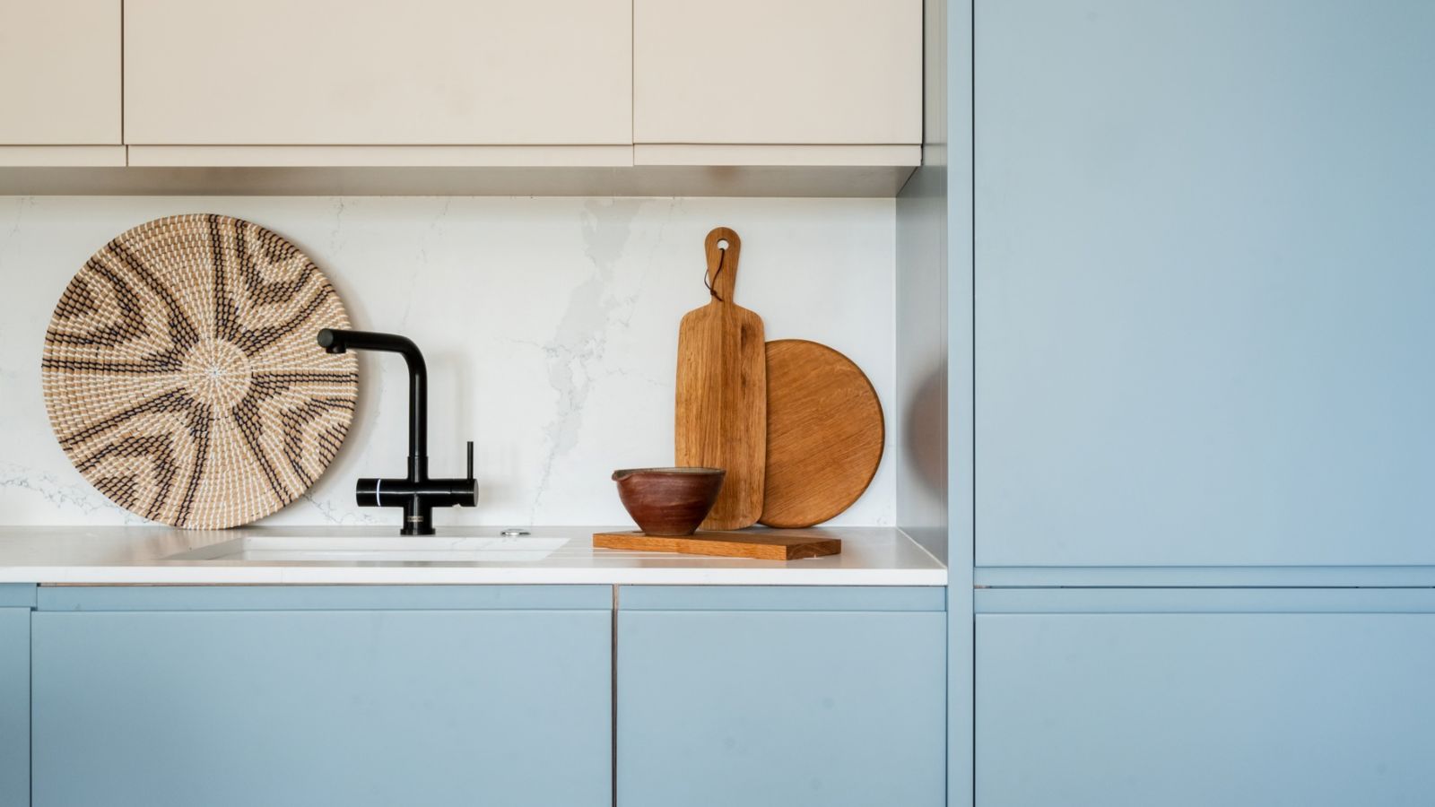 A modern kitchen with pale blue handleless base cabinets and cream upper units, featuring a white marble-effect splashback. A matte black angular tap sits above an undermount sink, with a round woven wall basket and wooden chopping boards and bowl used as decorative accessories on the white worktop.
