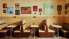 A restaurant banquette booth made of pale, camel-tinted leather, dark wood, and a table crafted with the same materials except for its chrome legs sits in a yellow-painted restaurant with stone floors, topped by two espresso cups, an open book, salt and pepper grinders in wood, a tiny flower vase, and a chrome table lamp.