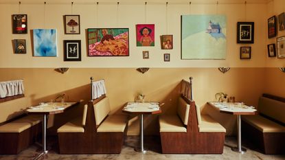 A restaurant banquette booth made of pale, camel-tinted leather, dark wood, and a table crafted with the same materials except for its chrome legs sits in a yellow-painted restaurant with stone floors, topped by two espresso cups, an open book, salt and pepper grinders in wood, a tiny flower vase, and a chrome table lamp.