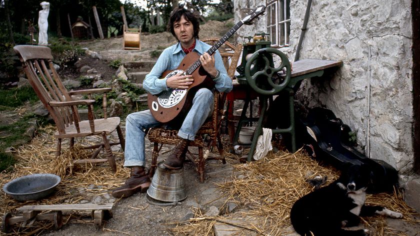 Ronnie Lane seated outside his country cottage, playing guitar 