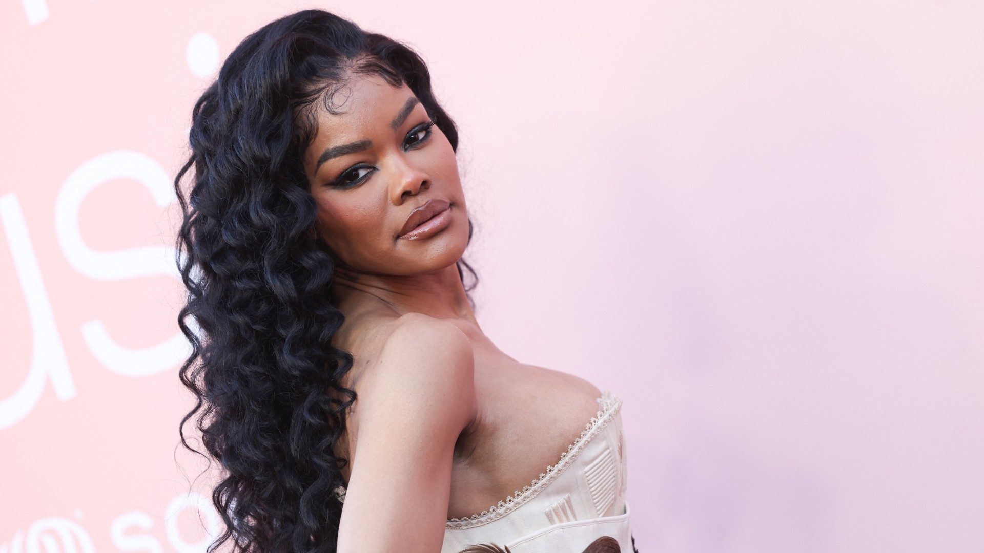 a black woman with long, curly hair poses in front of a pink backdrop at a red carpet event