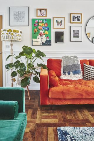 Living room with white panelled wall, parquet flooring, orange velvet sofa, green velvet sofa, gallery wall with colourful prints and a large green plant in a woven basket