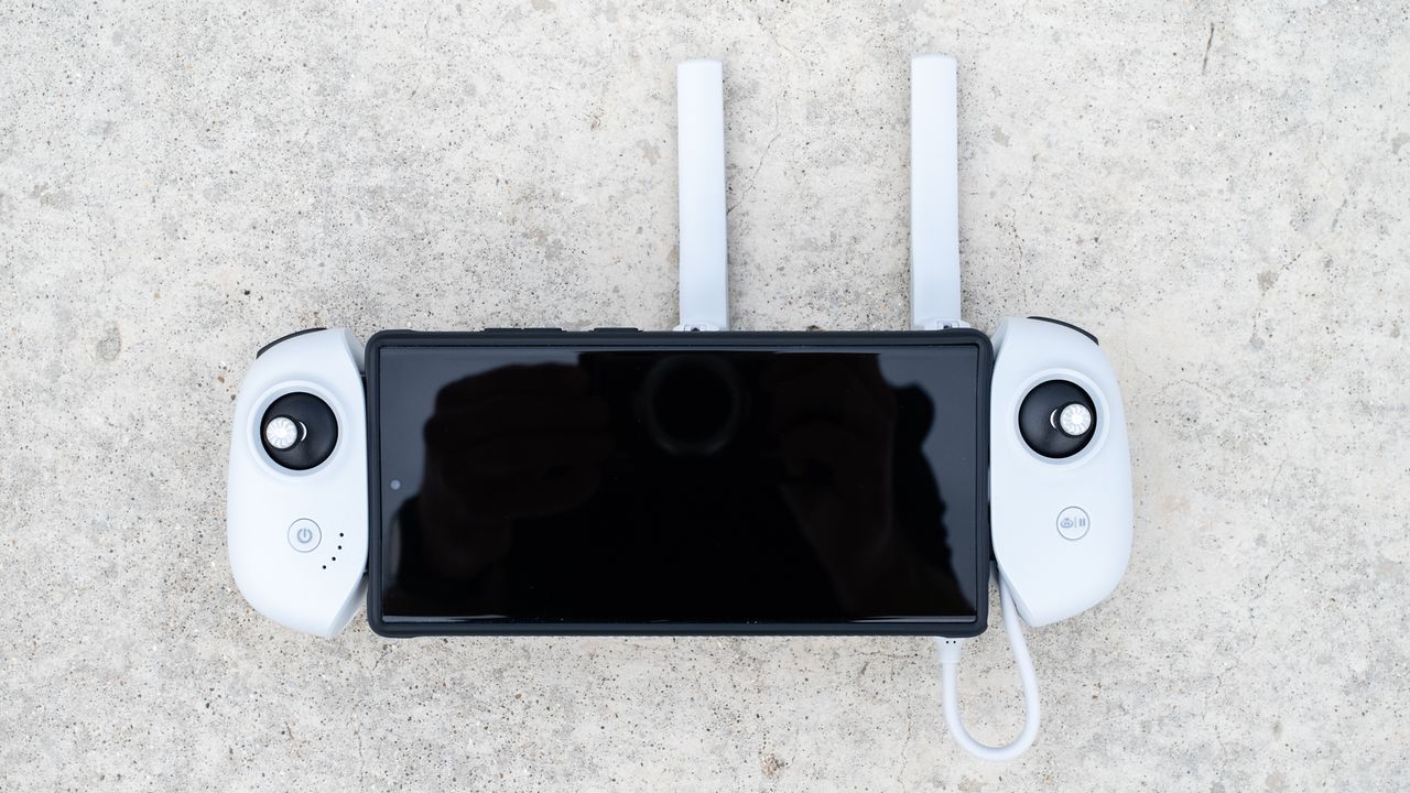the top-down view of a white controller device, with two back joysticks on the left and right near the top of the grips. A black-screened device is held between the white grips, with two antennas sticking upward.