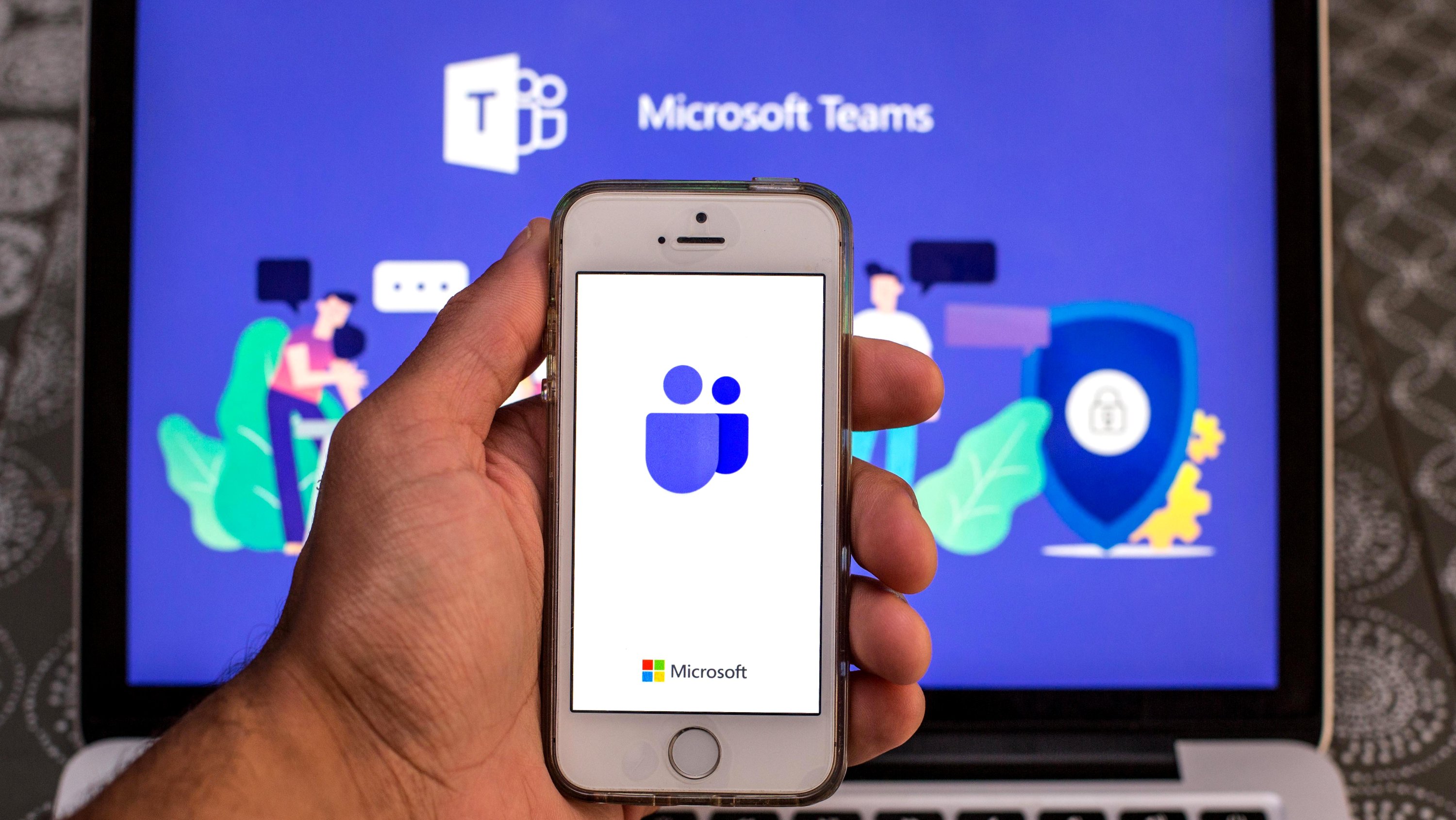 Microsoft Teams app seen displayed on a smartphone screen and a Microsoft Teams logo on a laptop in the background.