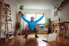 Man in sportswear in living room sits in a deep squat holding a resistance band in both hands above his head.