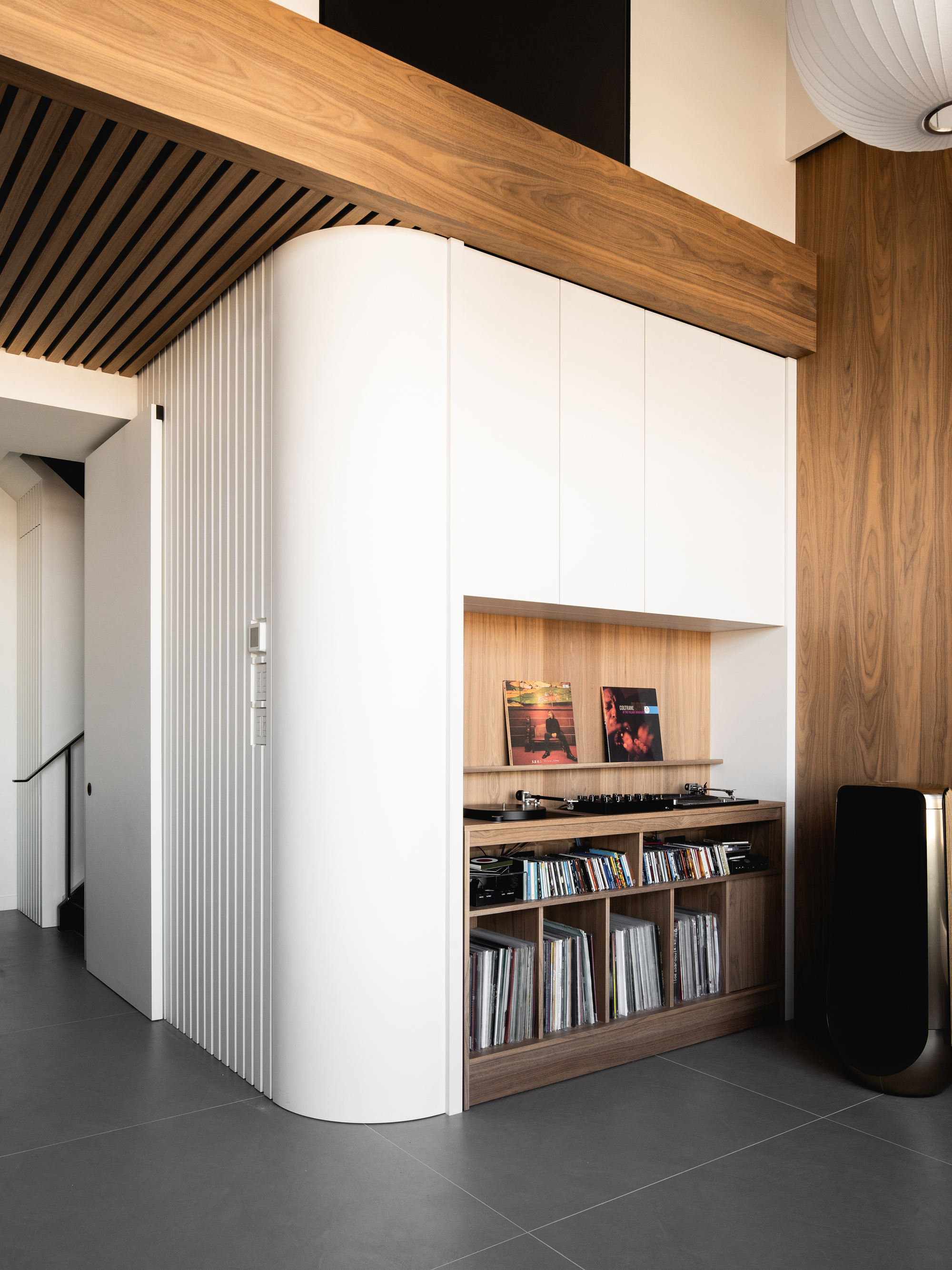 Image of a modern space with large gray tile floors, white walls, and wooden beams. The white wall has a wooden built-in vinyl bar with record storage, a vinyl player, and a shelf with vinyls styled on it.