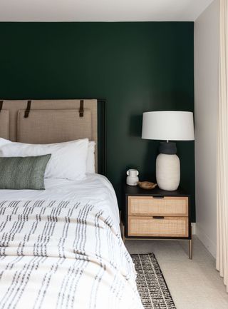 neutral bedroom scheme with forest green painted wall behind bed, black wood and wicker nightstand, white pendant, oatmeal upholstered headboard, white bedding, gray and white stripe blanket, gray cushion
