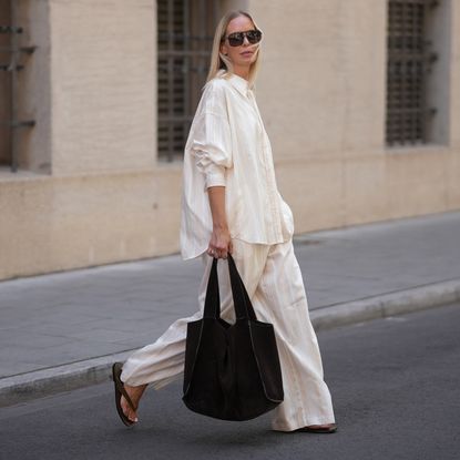 Mirja Klein is seen wearing a matching set consisting of a white striped oversized dobby-weave shirt with collar and buttons down in the front from H&M and a white striped straight pull-on trousers with loose fit and straight legs from H&M; a black suede tote bag with slouchy design and open top from COS; black oversized aviator sunglasses from Tom Ford; a pair of khaki green suede flip flops from A.EMERY on June 18, 2025 in Munich, Germany.