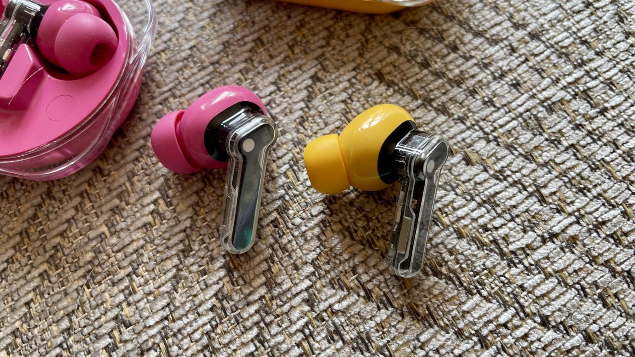 Nothing Ear (3a) in pink and Nothing Ear (a) in yellow, held in a hand and on beige background