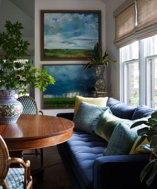seating area with dark navy blue sofa, round wooden table with large plant and landscape artwork on the walls