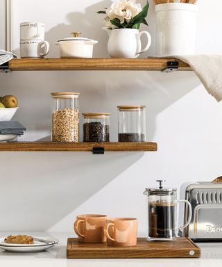 A white kitchen wall with two wooden shelves with white decor and flowers on the top one, glass canisters with grains and coffee grounds on the next, and a wooden chopping block with orange mugs and a French press on the counter