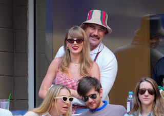 Taylor Swift wearing a red gingham dress and Travis Kelce wearing a striped bucket hat hugging her at the US Open