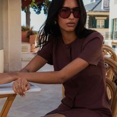 Model sitting on a wicker chair at a small stone-topped table outside in a button brown dress and big dark sunglasses