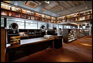 Interior view of Tsutaya Books featuring brown patterned carpet, windows, dark wood shelving units filled with records and an area with speakers and other music equipment