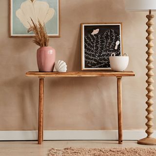a small wooden console table set against a brown earthy wall in a living room, with a wooden floor lamp with white lampshade next to it and a pink pot plant and black picture on top