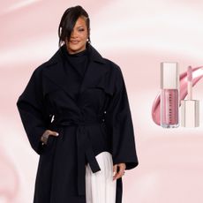 Rihanna posing against a pink backdrop wearing a black trench coat and white pants. There is a Fenty lip gloss, highlighter, and blush next to her.
