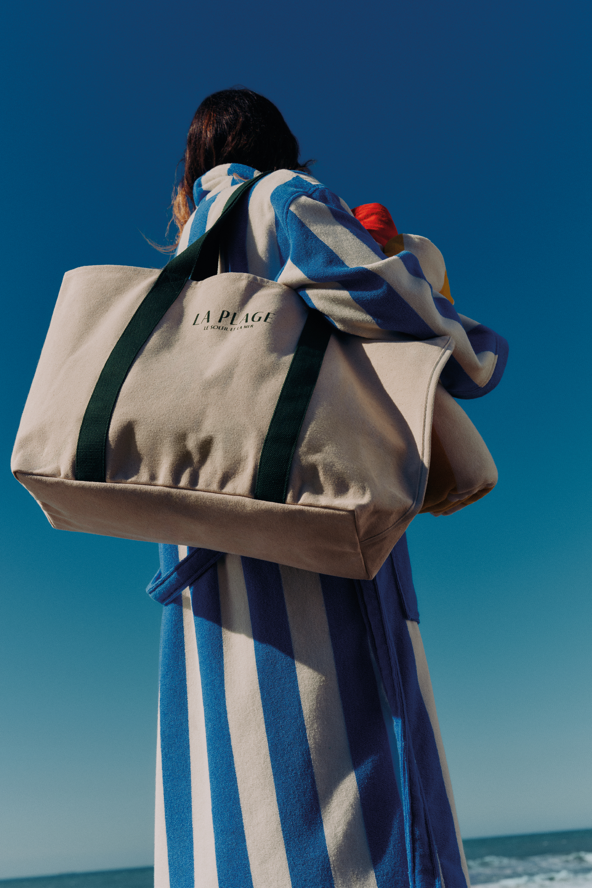 Image of a woman in a white and blue striped robe with a canvas bag over her arm standing with a view of blue skies and the sea in front of her.