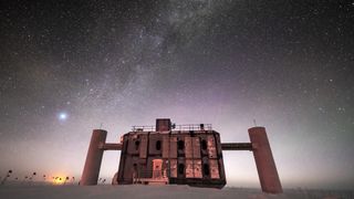 Front view of the IceCube Lab at twilight, with a starry sky showing a glimpse of the Milky Way overhead and sunlight lingerin​​g on the horizon.