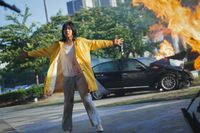 a Korean woman in a yellow coat and bloodstained clothes stands with flaming crashed cars behind her. 