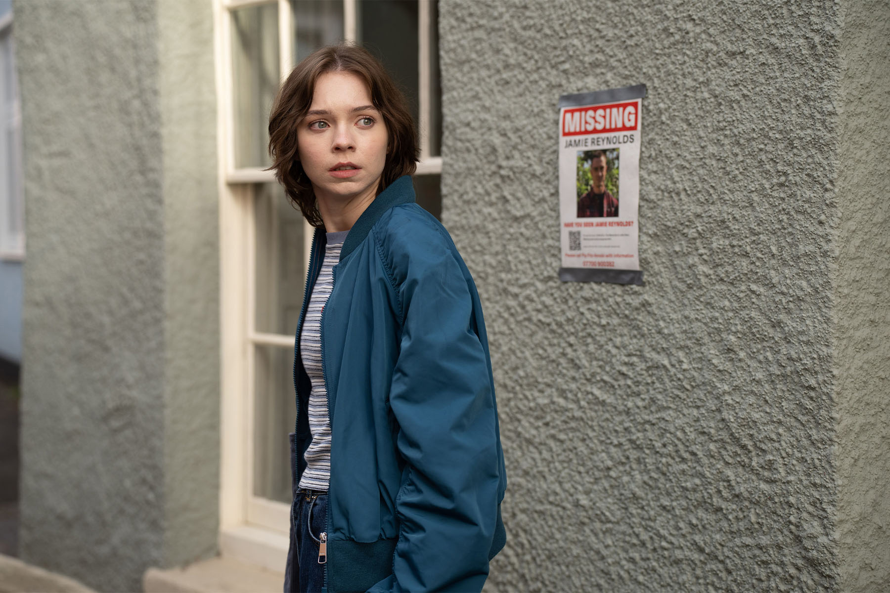 Emma Myers as Pip in season 2 of A Good Girl's Guide To Myrder. She is standing in front of a missing person sign wearing a blue windbreaker jacket, stripe t-shirt, and jeans.