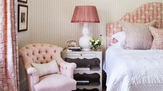 A white and pink bedroom with patterned wallpaper, a patterned headboard, white comforter, white lamp with pink lampshade, white nightstand, pink chair, patterned curtains, and framed artworks.