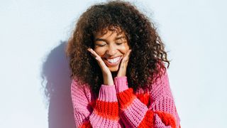 A young woman in a colorful jumper and with curly hair is seen caressing her face