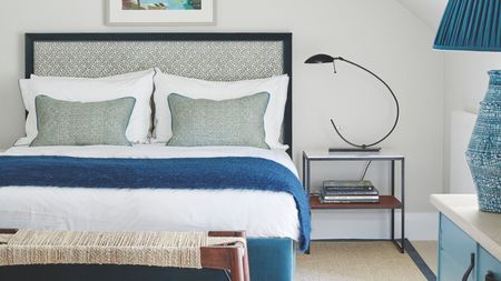 White and blue bedroom with a double bed with patterned headboard, white bedding, blue blankets and cushions, white and blue nightstand, blue lamp, rope and wood bench, and nightstand with curved lamp